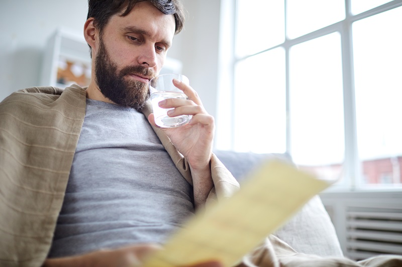 Young man wrapped into blanket drinking water and reading financial paper or letter while staying at home